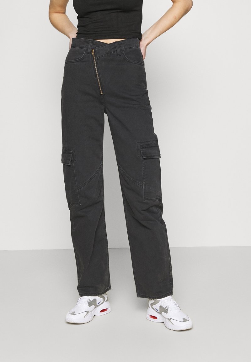 Personne portant un pantalon cargo noir taille haute avec une fermeture éclair frontale et des baskets blanches, debout, mains sur les hanches, sur un fond uni.