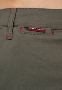 Pantalons en coton vert olive avec des accents de couture rouge, dotés d'une poche arrière avec une étiquette "Giro & Baxx". Texture lisse et coupe ajustée.