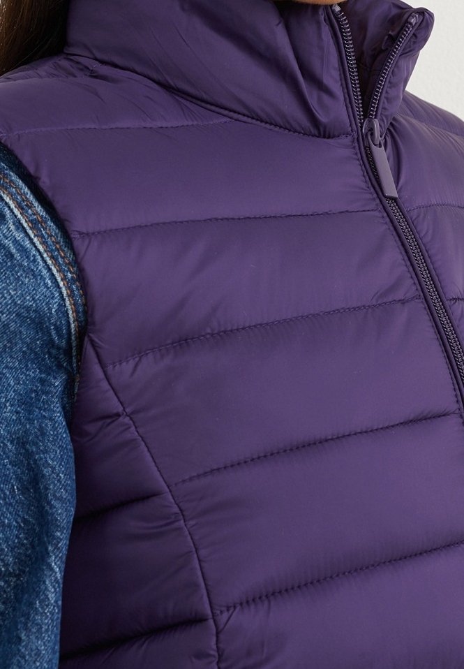 Gros plan d'un gilet matelassé violet porté sur une chemise en denim bleue, montrant la fermeture éclair avant et la zone du col.