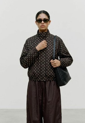 Femme portant des lunettes de soleil foncées et une veste marron à pois, tenant un sac à main noir tissé avec un pantalon marron foncé ample.