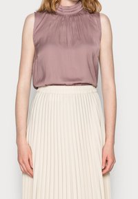 Halenka bez rukávů v popelavě růžové barvě s plisovanými detaily na výstřihu, v kombinaci se krémovou plisovanou sukní s elastickým pasem.