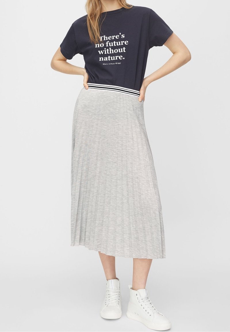 Femme debout, les mains sur les hanches, portant un t-shirt à slogan bleu marine, une jupe plissée gris clair et des baskets montantes blanches.