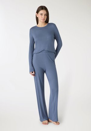 Młoda kobieta stojąca boso, ubrana w luźną, długotrwałą niebieską bluzkę z długim rękawem oraz pasujące szerokie spodnie, na tle jasnego, jednolitego tła.