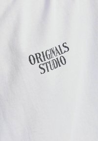 Camiseta de algodón blanca con un logo negro impreso que dice "ORIGINALS STUDIO" en una fuente gruesa y ligeramente desgastada.