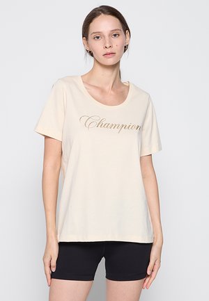 Donna con pelle chiara e capelli castani indossa una maglietta crema con la scritta "Champion" e pantaloncini da ciclista neri, in piedi davanti a uno sfondo semplice.