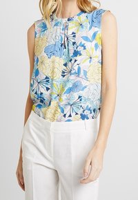 Blouse sans manches à motifs floraux en bleu, jaune et blanc. Présente un col rond et un pli subtil à l'avant. Assorti à un pantalon blanc.