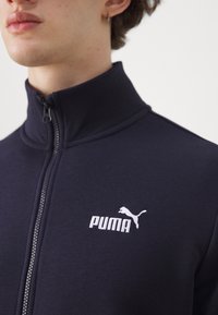 Marinblå zip-up jacka i mjukt material med hög krage, vitt Puma-logotyp och kontrasterande dragkedjedetaljer.