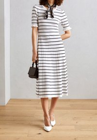 Frau in einem weißen Midikleid mit schwarzen horizontalen Streifen, weißen Absätzen, einem schwarzen Halstuch und einer kleinen schwarzen Handtasche.