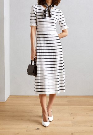 Mujer vestida con un vestido midi blanco con rayas horizontales negras, tacones blancos, bufanda negra en el cuello y sosteniendo un pequeño bolso negro.