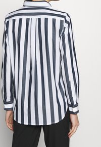 Button-up skjorta i bomull med marinblå och vita vertikala ränder, långa ärmar, krage och rundad nederkant; har knäppta manschetter och knappslå.