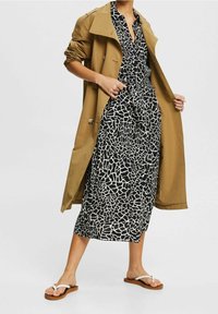 Béžový trench coat přes černobíle vzorované šaty s designem do zavinovacího stylu, doplněný bílými žabkami s hnědou podrážkou.