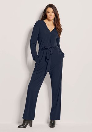 Marineblauer Overall mit langen Ärmeln, V-Ausschnitt und Taillengürtel. Hergestellt aus glattem Stoff, verfügt über seitliche Taschen und ein weites Bein-Design, kombiniert mit schwarzen High Heels.