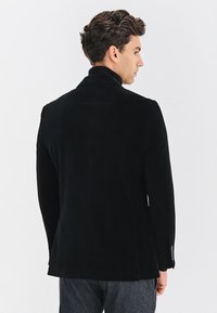 Mężczyzna z kręconymi włosami, ubrany w czarną marynarkę i ciemne spodnie, stoi lekko odwrócony w stronę białego tła.