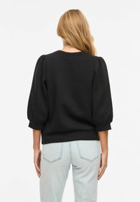 Femme aux longs cheveux blonds portant un haut noir à manches bouffantes et un jean bleu clair, vue de dos sur un fond blanc uni.