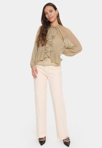 Light beige sheer blouse with ruffle detailing at the front, long sleeves, and a tie at the neck, paired with cream wide-leg trousers and black heels.