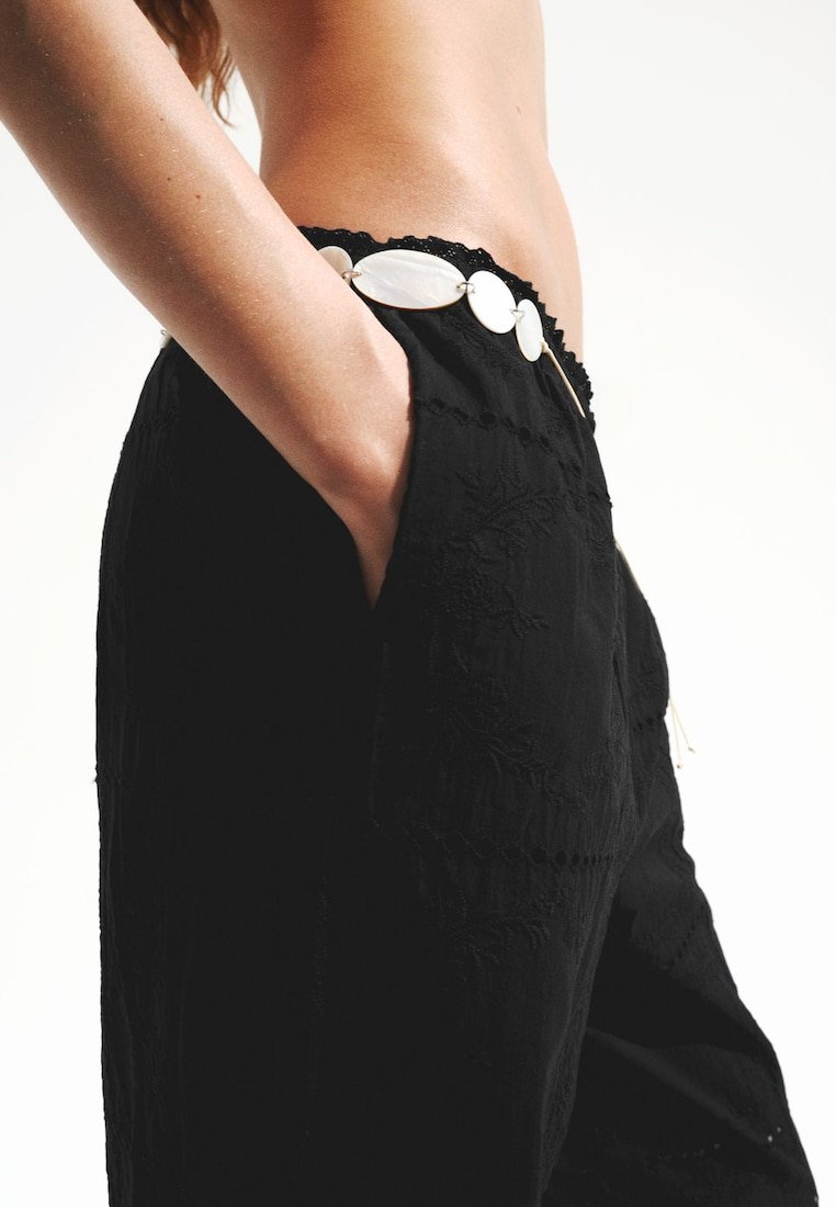 Femme portant un pantalon noir brodé avec une ceinture décorative blanche, main placée dans la poche, vue de la taille vers le bas.