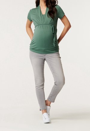 Těhotenský top v zelené látce s řaseným pasem a detailem vázání, kombinovaný s světle šedými skinny džíny a bílými teniskami.