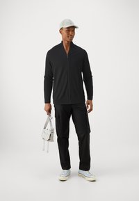 Giacca nera con zip, maniche lunghe, indossata con pantaloni cargo neri. Sneakers azzurre chiare e un cappellino grigio chiaro. Tiene in mano una piccola borsa grigia chiara.