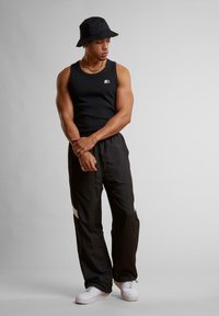 Svart tanktop, svarte joggebukser med hvite detaljer, og en svart bøttehatt; modellen har på seg hvite sneakers; atletisk kroppsbygning.