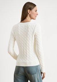 Ženska v profilu, oblečena v bel pulover z vzorcem pletenice in modre kavbojke, z ravnimi rjavimi lasmi, ki so ji potisnjeni za uho.