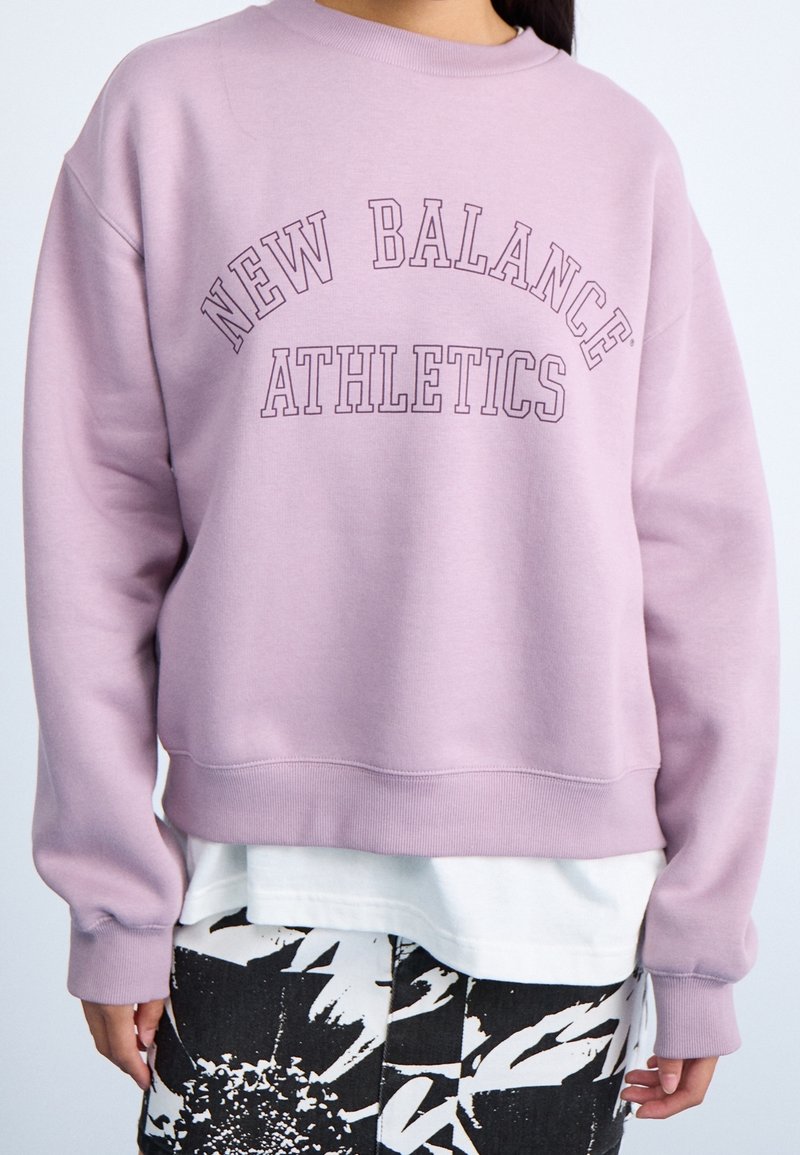 Sudadera de color púrpura claro con cuello redondo, con el texto "NEW BALANCE ATHLETICS" en un tono púrpura más oscuro. Puños y dobladillo acanalados.