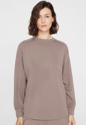 Femme aux cheveux courts et bruns portant un haut ample à manches longues taupe avec des rayures côtelées sur les manches, sur fond blanc.