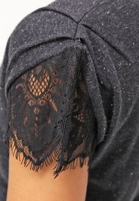 Haut en tricot noir avec des manches courtes et une délicate bordure en dentelle noire, mettant en valeur des motifs complexes et une finition à franges pour un détail supplémentaire.