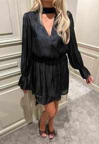 Rochie neagră, cu mâneci lungi, realizată dintr-un material mătăsos. Are un decolteu în formă de V și o talie strânsă. Este asortată cu tocuri deschise la vârf, cu un model.