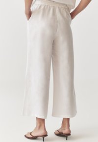 Pantalons larges en lin beige clair avec une taille élastique, des poches latérales et une texture lisse, associés à des sandales à talons minimalistes.