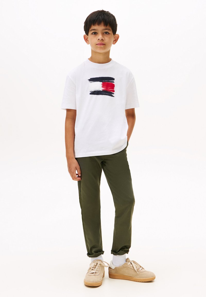 T-shirt blanche à manches courtes avec un logo à coups de pinceau bleu et rouge, assortie à un pantalon vert olive et des baskets beiges avec des accents blancs.