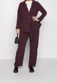 Traje burdeos de dos piezas que incluye una chaqueta con cinturón, solapas y bolsillos laterales, combinada con pantalones de corte holgado y zapatos negros.