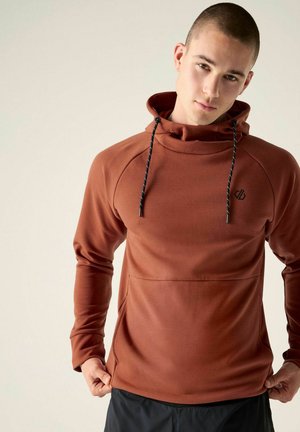 Young man with short hair wearing a rust-colored hoodie with black drawstrings and black pants against a plain light background.