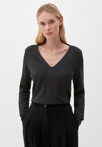 Schwarzes Langarmoberteil mit V-Ausschnitt, das eine figurbetonte Passform und dezente Nahtdetails aufweist. Kombiniert mit einer hoch geschnittenen schwarzen Hose.