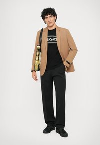 Man in een tan blazer, zwart Versace T-shirt, zwarte broek, zwarte schoenen en met een zwart en goud patroon tote bag op de schouder.