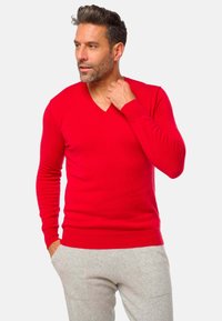 Pull rouge en V en tricot épais, à manches longues et avec des poignets côtelés, porté sur un pantalon de survêtement gris sur un fond neutre.