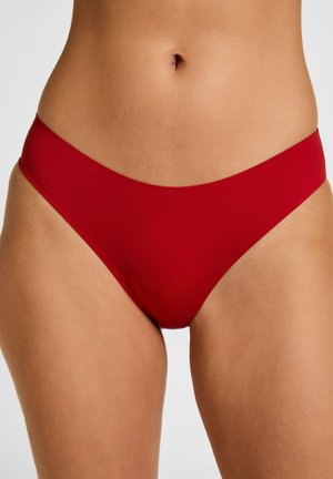 Middenstuk van een vrouw die naadloze rode bikini-onderkleding draagt tegen een effen achtergrond.