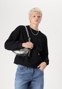Sudadera negra con hombros caídos, jeans azules y un bolso bandolera metálico plateado con forma curva y correa ajustable.