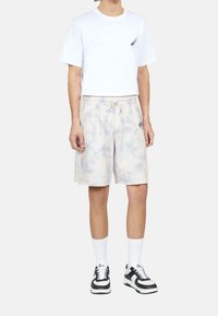 Vit t-shirt med kort ärm och en fjäderlogo, tillsammans med krämfärgade tie-dye-shorts med elastisk midja. Svarta och vita sneakers.