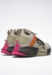 Un par de zapatillas Reebok en beige y negro con suelas gruesas, detalles en el talón de color rosa, cintas laterales naranjas y detalles del logo, vistas desde la parte trasera.