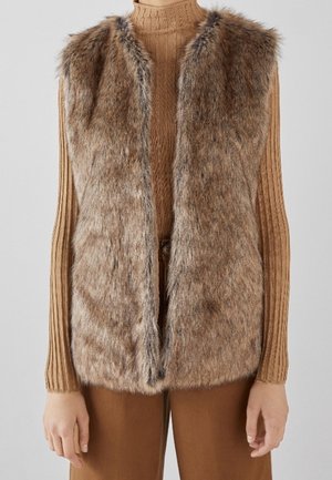Braune Kunstpelzweste über einem gerippten Kamelrollkragenpullover und braunen Hosen, von den Schultern bis zu den Hüften gezeigt.