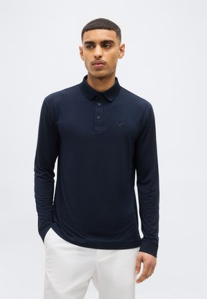 Noor mees lühikeste juustega, kandes tumelise sinist pika varrukaga polosärki ja valgeid pükse, seisab käed taskus, neutraalne taust.