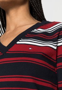 Pull en tricot rayé bleu marine, rouge et blanc. Design en V avec un petit logo. Texture douce avec des rayures larges et étroites alternées.