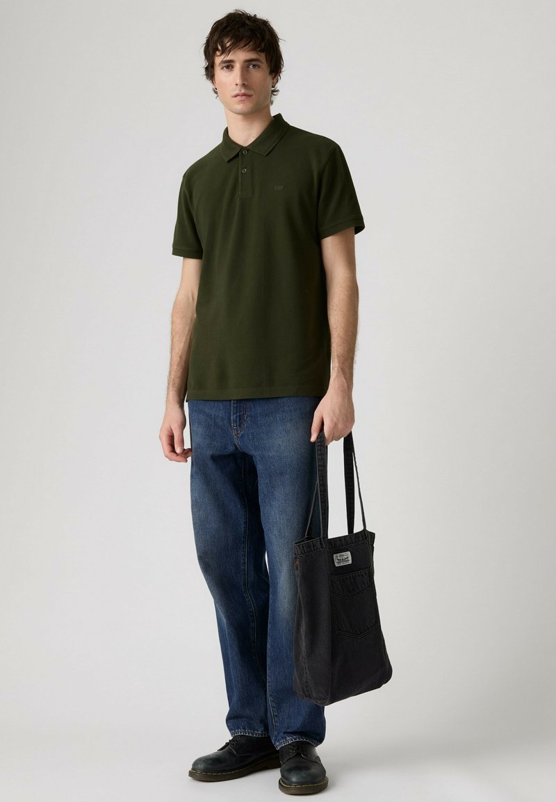Jeune homme portant un polo vert olive, un jean bleu, des chaussures noires, tenant un sac fourre-tout noir sur un fond uni.