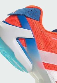 Detailní záběr na červenou a modrou atletickou obuv s bílou podešví a světle modrým vzorkem, která má prodyšný síťovaný svršek a podpůrné strukturální prvky.