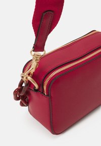 Sac à main en cuir rouge avec une finition texturée, des zips dorés et un accent en chaîne. Il dispose d'une bandoulière en tissu amovible pour plus de polyvalence.
