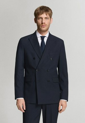 Hombre vestido con un traje oscuro de doble botonadura, camisa blanca y corbata azul marino, posando frente a un fondo gris claro liso.