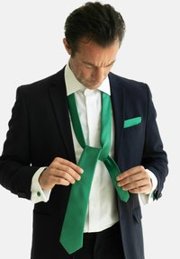 Žalias šilkinis kaklaraištis, pakabintas ant baltos marškininės, dėvimas su juodu švarku. Kaklaraištis turi lygų paviršių ir surištas laisvu mazgu.