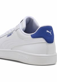 Sneaker Puma bianco con accenti blu, parte superiore in sintetico liscio, linguetta sul tallone testurizzata e suola piatta in gomma. Presenta il marchio sul lato e sul tallone.