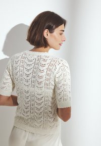 Mujer con cabello corto y castaño, vestida con un suéter blanco de punto de manga corta y una falda de color claro, de pie contra una pared blanca lisa.