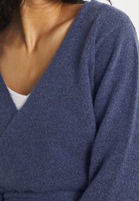 Blauer gestrickter Pullover mit tiefem V-Ausschnitt, strukturiertem Stoff und weiten Ärmeln. Über einem weißen Oberteil getragen, was einen gemütlichen Look kreiert.
